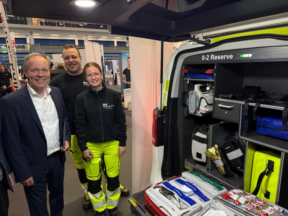Drei Personen stehen neben einem geöffneten Rettungswagen mit sichtbarer medizinischer Ausrüstung. Im Hintergrund sind Messe- oder Ausstellungsstände erkennbar.