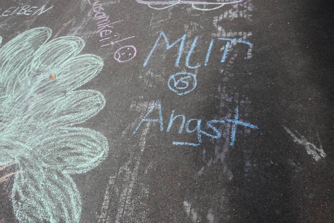 Mit Kreide auf Asphalt gemalte Wörter und Zeichnungen. Zu sehen sind die Wörter 'Mut vs Angst' und 'Einsamkeit' sowie ein großes Blumenmotiv.