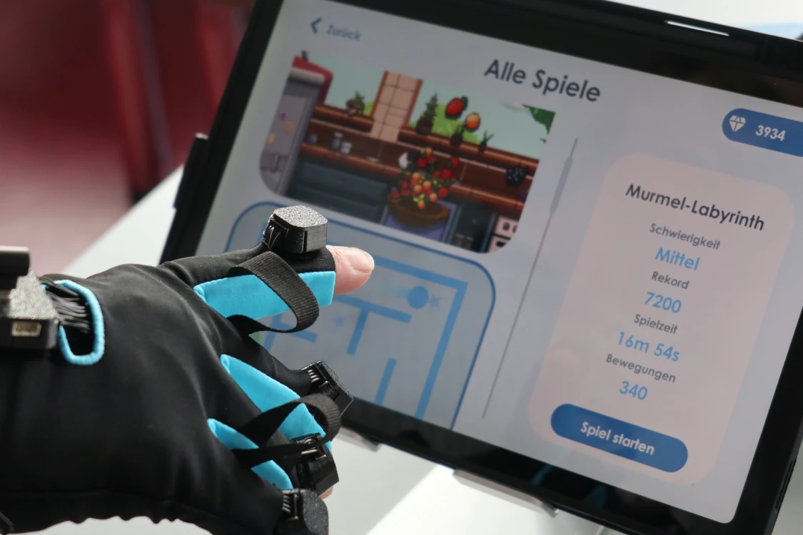 Eine Hand trägt einen schwarzen Handschuh mit blauen Akzenten und Sensoren, die auf die Finger montiert sind. Die Hand berührt einen Touchscreen, der ein Spielmenü mit dem Titel 'Alle Spiele' und Details zu einem Spiel namens 'Murmel-Labyrinth' anzeigt.