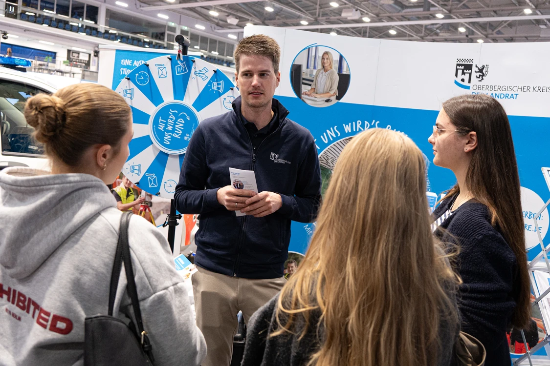 Ein Mann in dunkelblauer Jacke spricht mit drei jungen Frauen vor einem Messestand. Im Hintergrund ein Glücksrad und ein Plakat mit Logo und Text.