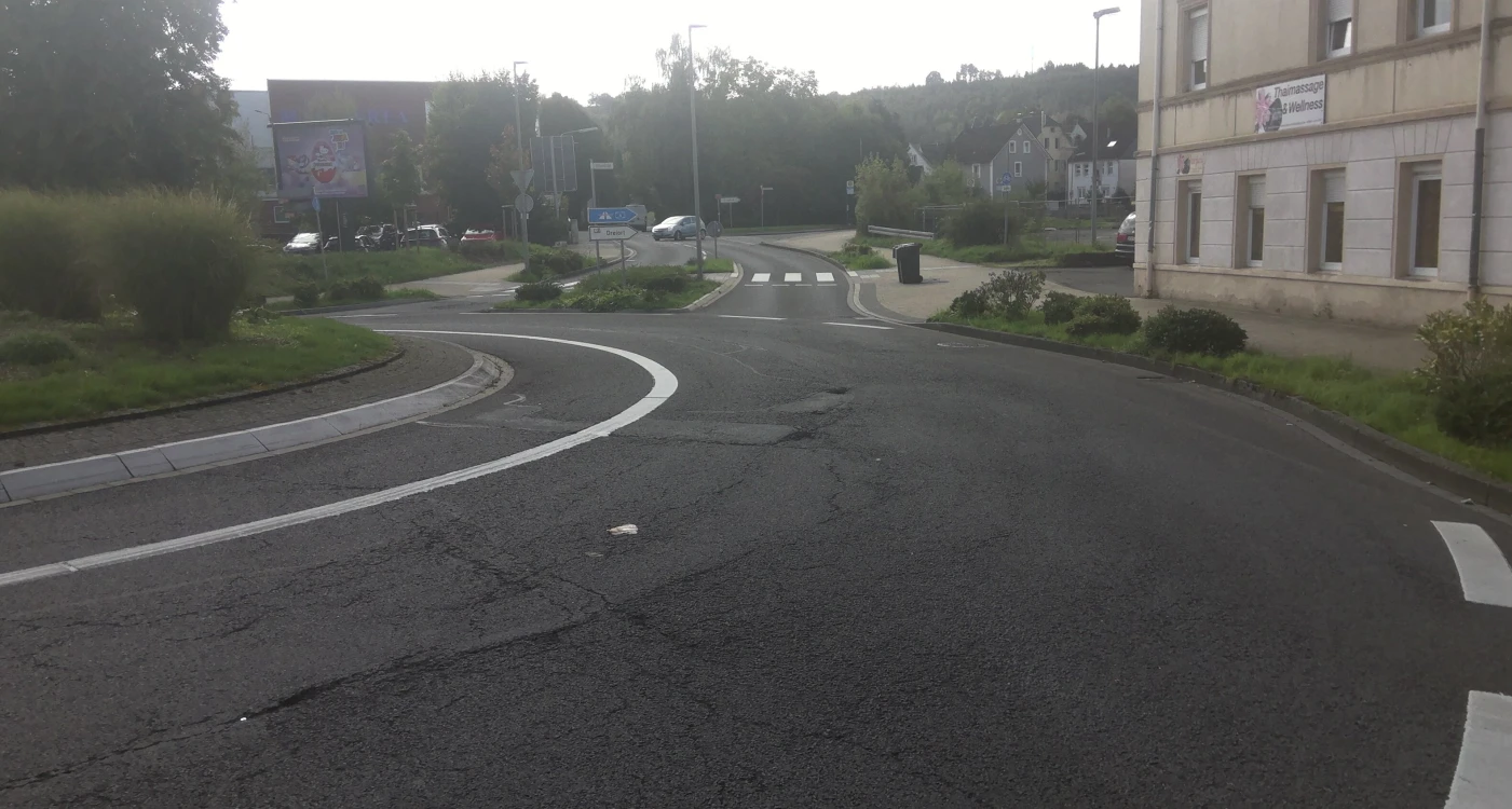 Straßenkreuzung mit Kreisverkehr, Zebrastreifen, umliegendem Grün und Gebäuden. Links eine Werbetafel und rechts ein Gebäude mit Wellness-Schriftzug.