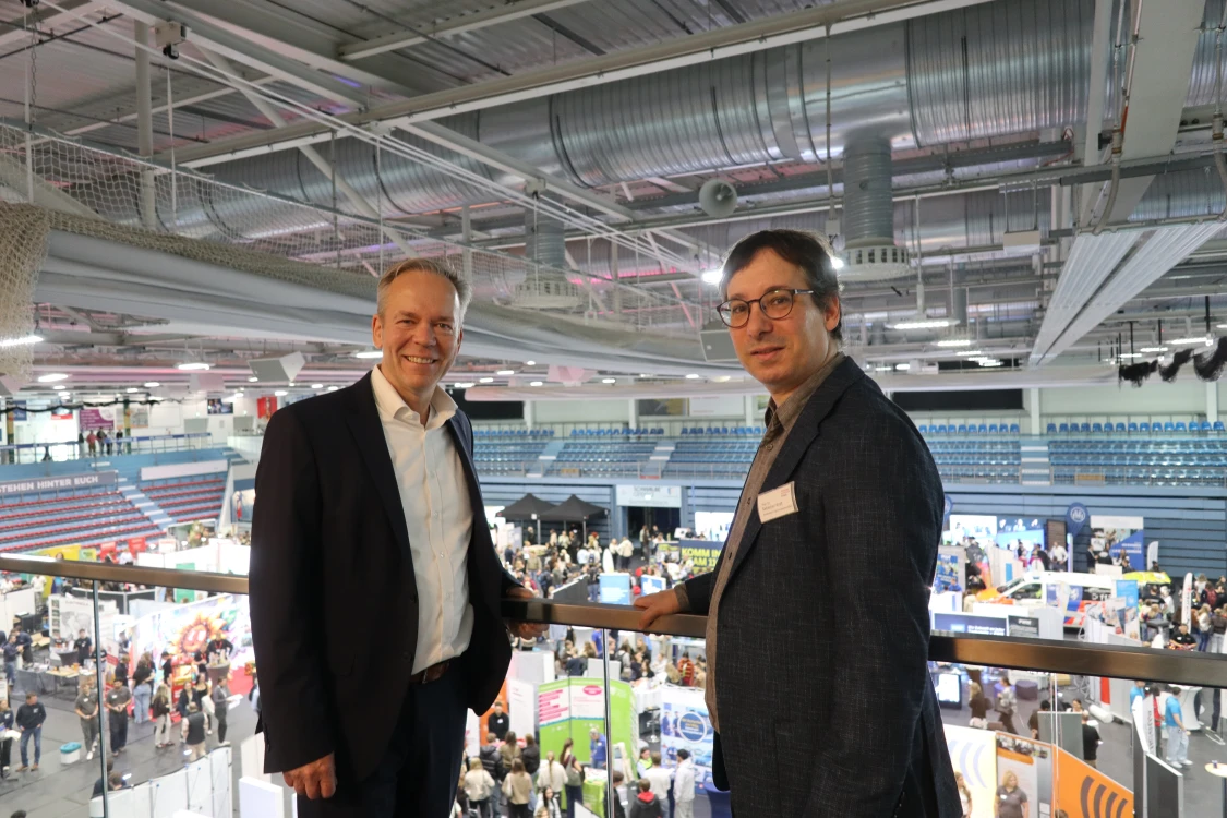 Zwei Männer stehen auf einer Galerie in einer großen Halle mit mehreren Ausstellungsständen. Im Hintergrund sind Menschen und Sitzreihen zu sehen.