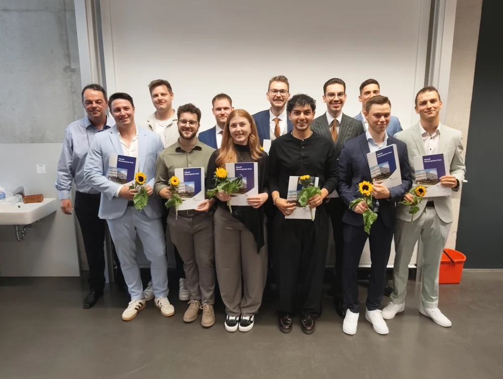Gruppenfoto von Personen, die Auszeichnungen und Sonnenblumen halten, in einem Innenraum mit weißer Wand und Waschbecken im Hintergrund.