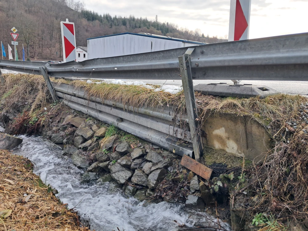 Eine beschädigte Leitplanke an einer Straße neben einem kleinen Bach. Die Böschung ist mit Steinen befestigt, einige Pflanzen wachsen am Ufer. Im Hintergrund sind ein Gebäude und ein bewaldeter Hügel zu sehen.