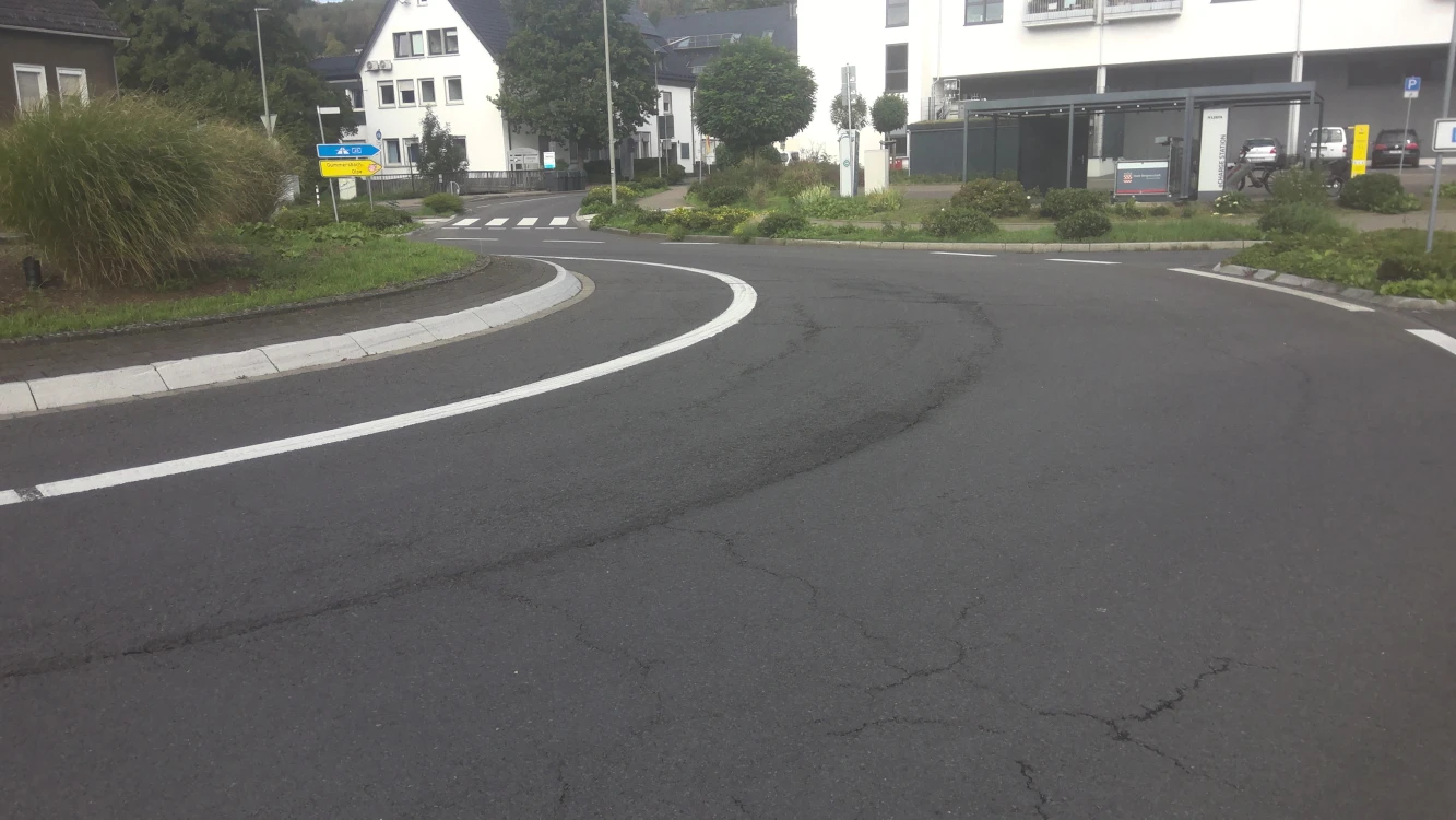Eine asphaltierte Straße mit einer Kurve, Straßenmarkierungen und einem begrünten Mittelstreifen. Im Hintergrund Gebäude, Schilder und ein Parkplatz sichtbar.