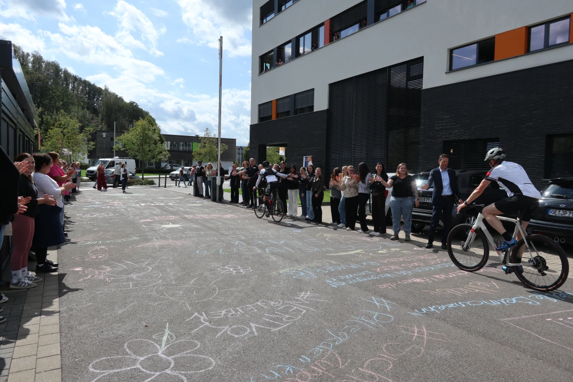 Eine Straße mit bunter Kreidemalerei, gesäumt von applaudierenden Menschen. Zwei Radfahrer fahren durch die Mitte. Moderne Gebäude im Hintergrund.