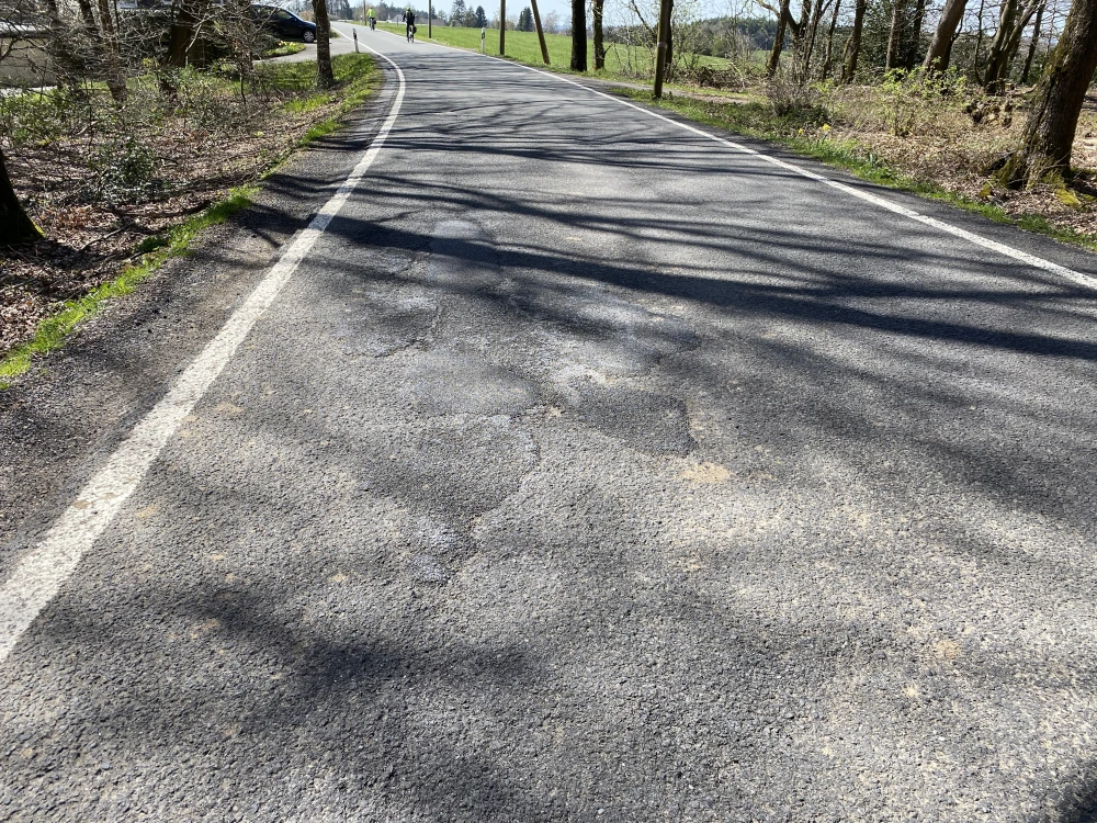 Eine asphaltierte Straße mit sichtbaren Schlaglöchern und Rissen, umgeben von Bäumen und Gräsern. Im Hintergrund sind Personen und Fahrzeuge erkennbar.