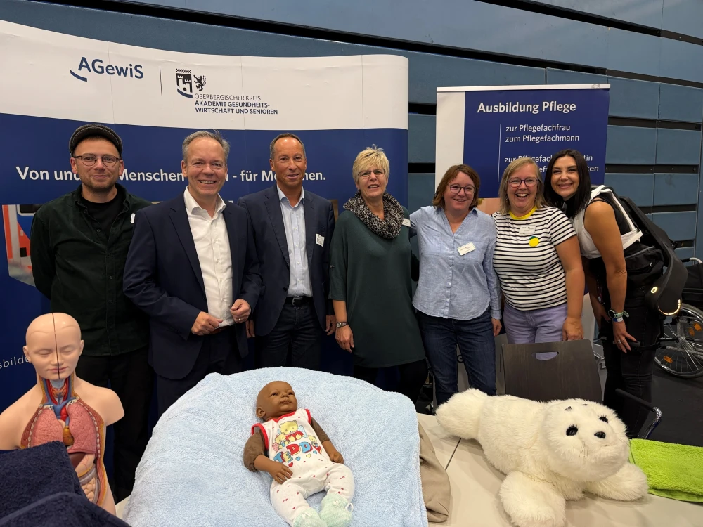 Eine Gruppe von sieben Personen steht vor Infotafeln zu Ausbildung und Pflege. Davor sind ein Anatomiemodell, eine Babypuppe und ein Plüschtier zu sehen.