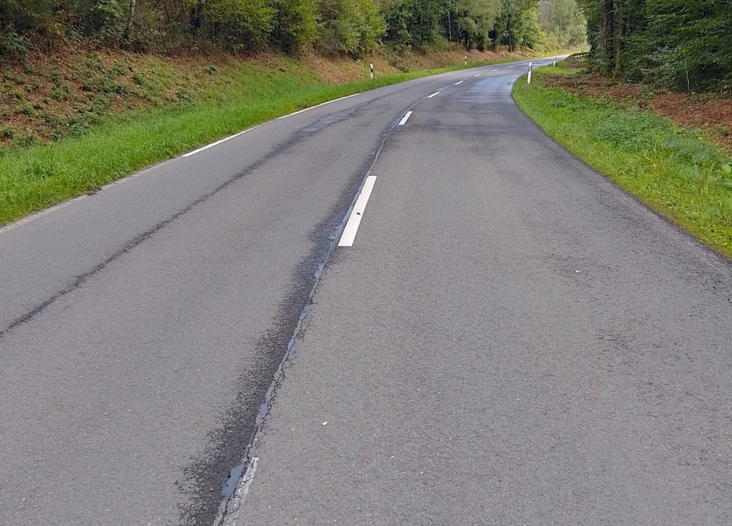 Eine asphaltierte Landstraße mit einer weißen Mittellinie, umgeben von grünen Wiesen und Bäumen. Der Himmel ist teilweise sichtbar und wirkt bewölkt.