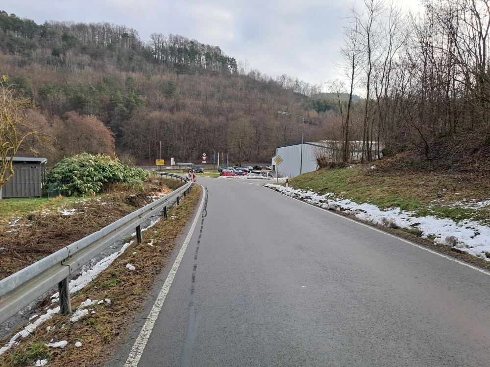 Eine asphaltierte Straße mit Leitplanke führt bergab in eine Kurve. Links und rechts sind schneebedeckte Grasflächen und Bäume. Im Hintergrund stehen Autos und ein Gebäude vor einem bewaldeten Hügel.