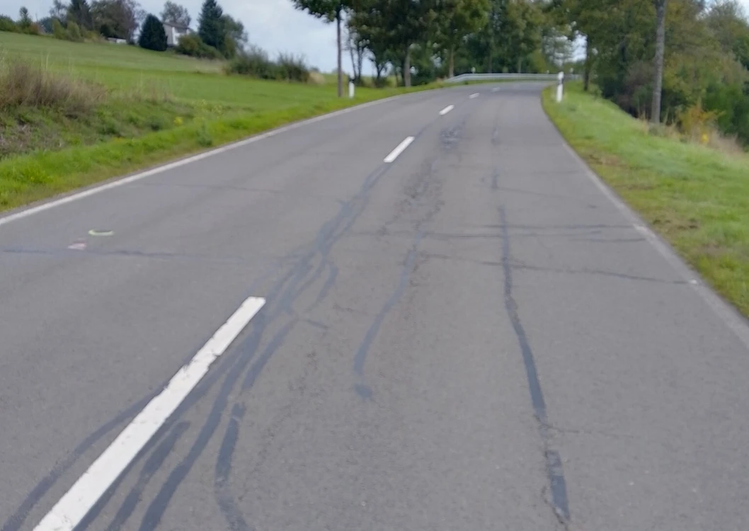 Eine asphaltierte Landstraße mit weißen Markierungen und Reparaturspuren, die durch eine grüne Landschaft mit vereinzelten Bäumen und Häusern führt. Der Himmel ist bewölkt.