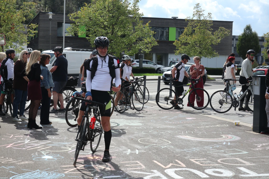 Eine Gruppe von Menschen, darunter Radfahrer in Sportkleidung mit Fahrrädern, steht auf einem Platz mit Kreidezeichnungen. Im Hintergrund sind Bäume, ein Gebäude und parkende Autos zu sehen.
