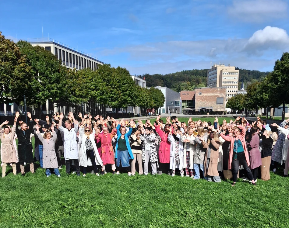 Eine große Gruppe von Menschen steht auf einer Wiese vor modernen Gebäuden. Sie tragen überwiegend Bademäntel und heben ihre Arme in die Luft. Im Hintergrund sind Bäume und ein Hügel zu sehen.