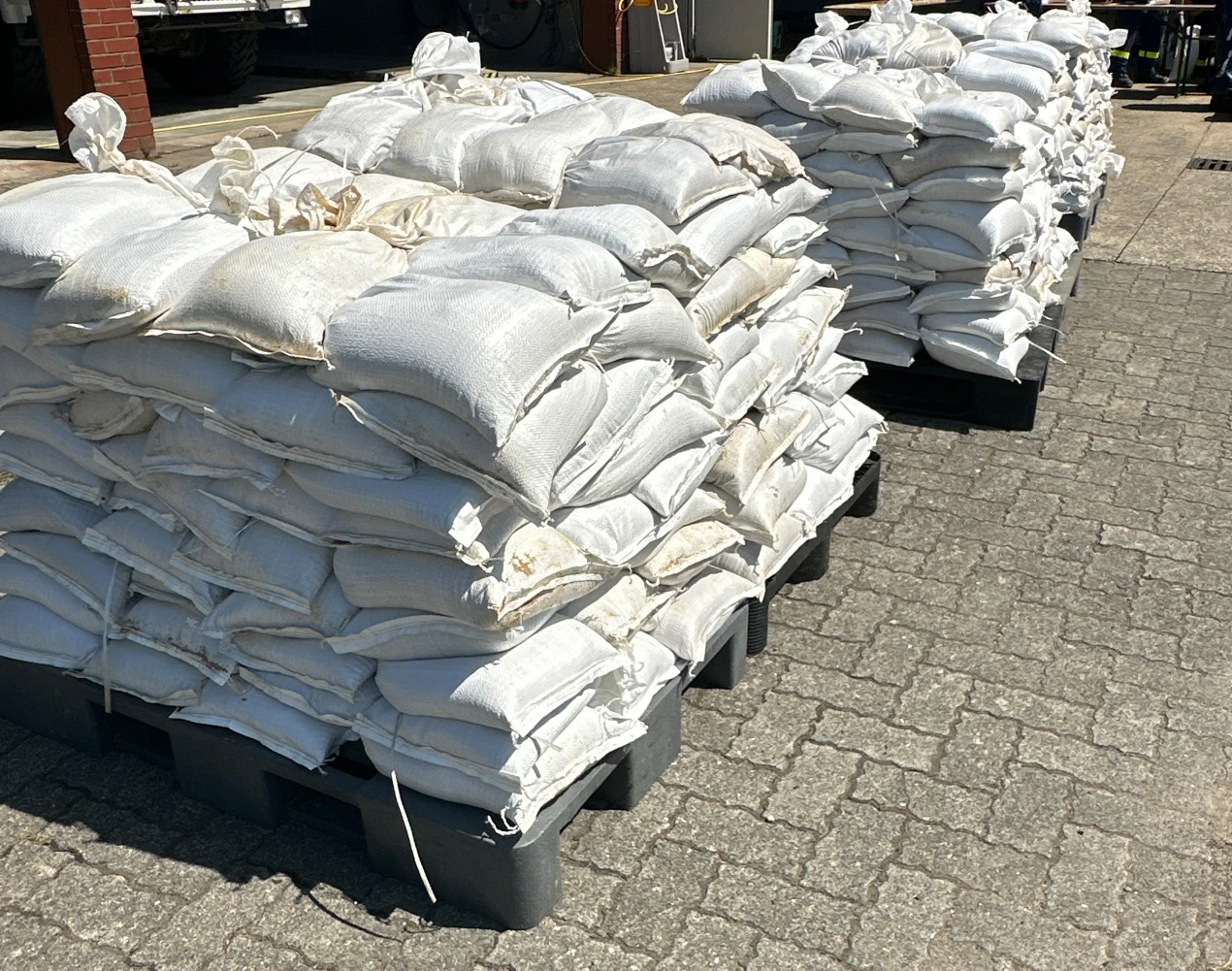 Mehrere Stapel von gefüllten Sandsäcken, die auf schwarzen Paletten auf einem gepflasterten Boden gelagert sind. Im Hintergrund sind Teile eines Gebäudes und Fahrzeuge sichtbar.
