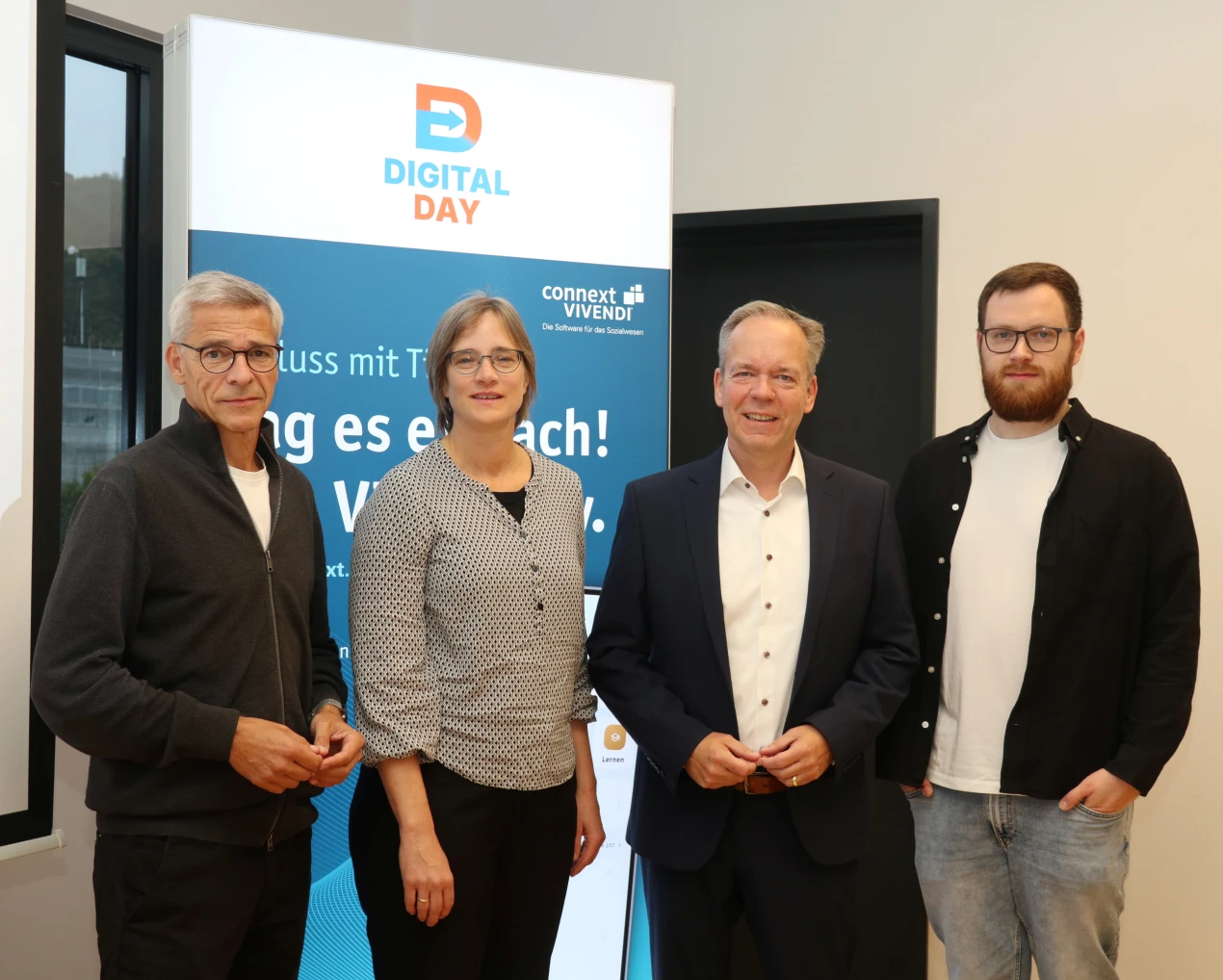 Vier Personen stehen vor einem Roll-Up mit der Aufschrift 'Digital Day' und weiteren Texten. Zwei Männer und eine Frau tragen Businesskleidung, ein weiterer Mann ist lässig gekleidet.