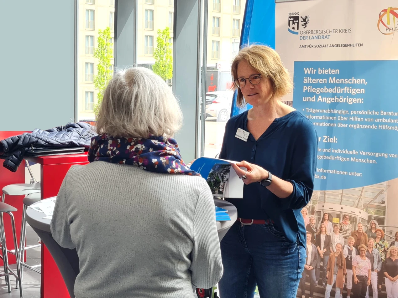 Zwei Frauen stehen an einem Stehtisch in einem Innenraum. Eine Frau mit Brille hält Broschüren in der Hand, während die andere mit dem Rücken zur Kamera steht. Im Hintergrund ist ein blaues Banner mit Text und ein großes Fenster sichtbar.