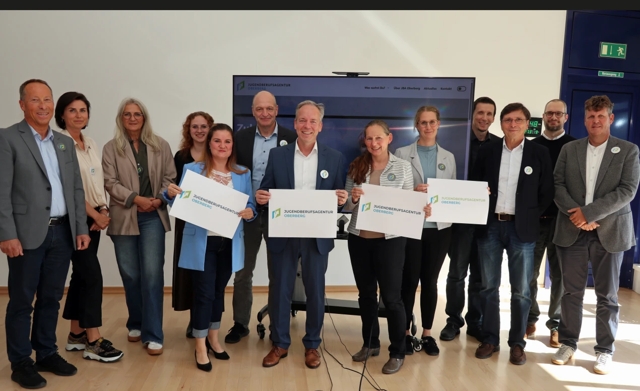 Gruppenfoto von 13 Personen in einem hellen Raum. Einige halten Schilder mit der Aufschrift 'Jugendberufsagentur Oberberg'. Im Hintergrund ein Bildschirm und eine Tür.