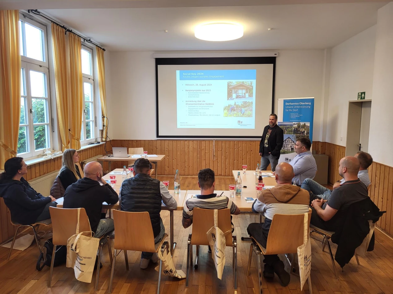 Ein Seminarraum mit mehreren Personen, die um einen Tisch sitzen und einem Vortragenden zuhören. Im Hintergrund eine Präsentation auf einer Leinwand.