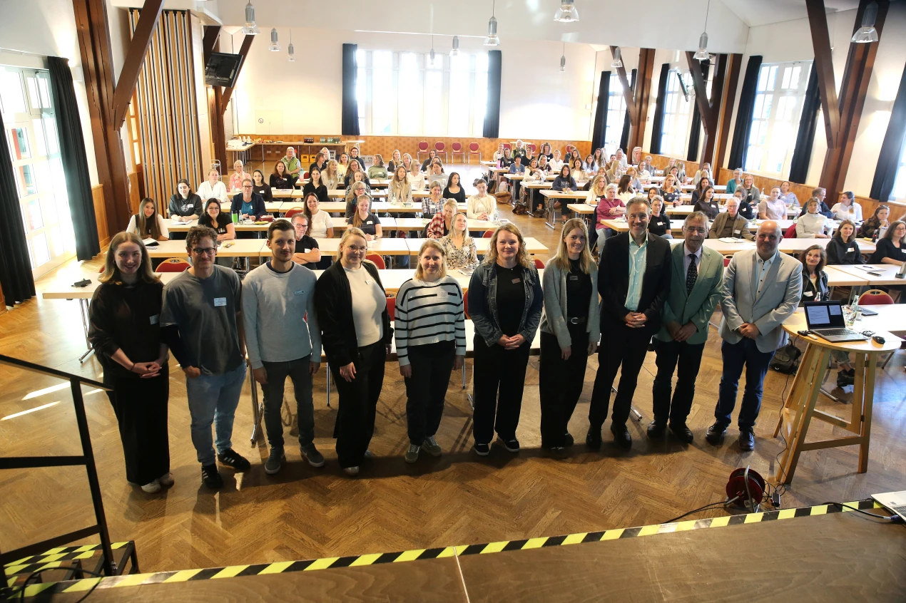 Eine Gruppe von Personen steht in einer Reihe vor einem Publikum in einem großen, gut beleuchteten Raum mit Holzboden und hohen Decken. Im Hintergrund sitzen Menschen an Tischen, die auf die Bühne blicken.