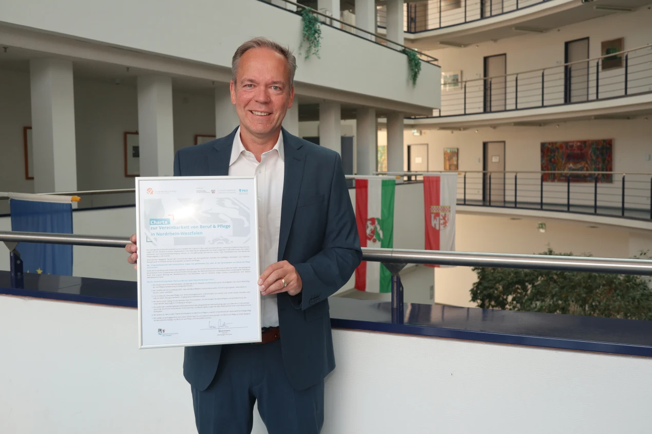 Eine Person in einem Anzug hält ein gerahmtes Dokument mit der Aufschrift 'Charta zur Vereinbarkeit von Beruf & Pflege in Nordrhein-Westfalen'. Im Hintergrund ist ein Innenbereich mit Geländern, Pflanzen und Flaggen zu sehen.