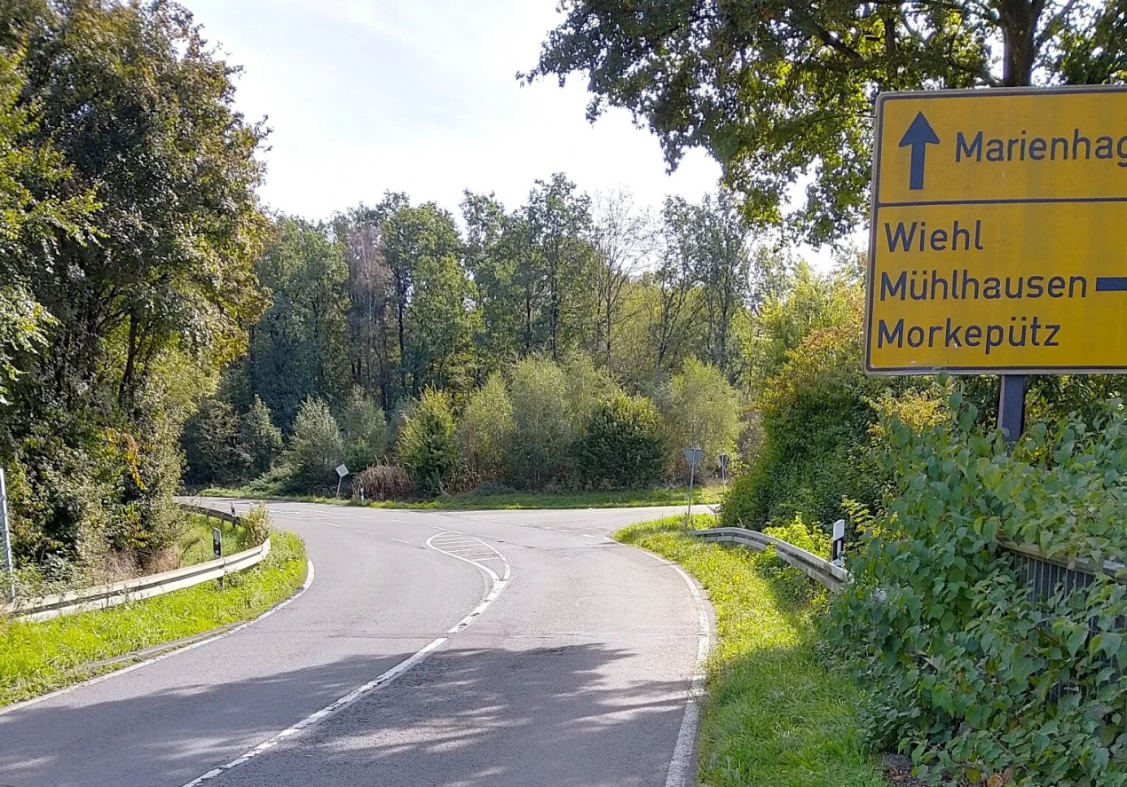 Straßenkreuzung mit Beschilderung. Gelbes Schild zeigt Richtungen nach Marienhagen, Wiehl, Mühlhausen und Morkepütz. Umgeben von grüner Vegetation und Bäumen.