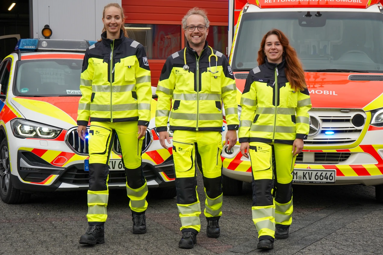 Drei Personen in leuchtend gelber Rettungsdienstkleidung stehen vor zwei Einsatzfahrzeugen mit rot-gelbem Design und eingeschaltetem Blaulicht.