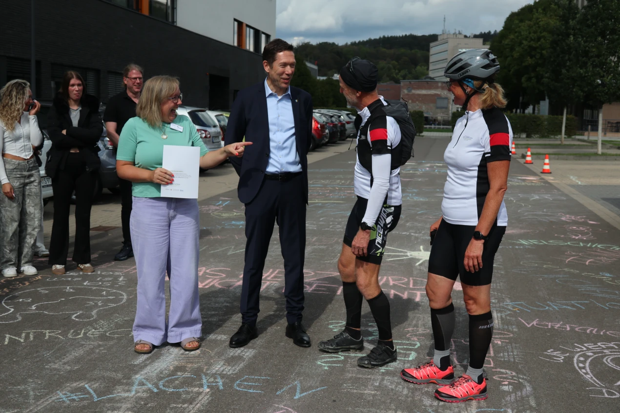 Eine Gruppe von Menschen steht auf einer mit bunter Kreide bemalten Straße. Eine Frau mit grüner Bluse hält ein Dokument und spricht mit einem Mann im Anzug. Zwei Personen in Fahrradkleidung stehen daneben. Im Hintergrund sind geparkte Autos und ein Gebäude zu sehen.