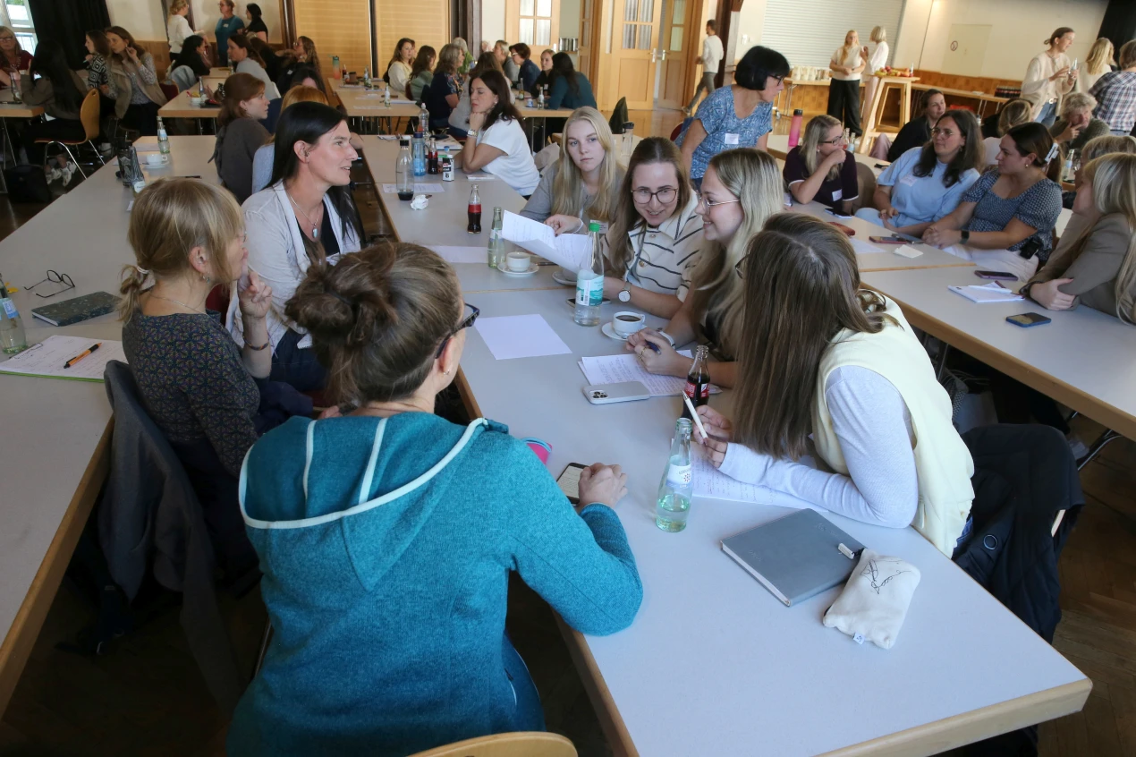 Eine Gruppe von Menschen sitzt an Tischen in einem Raum und diskutiert. Auf den Tischen liegen Notizen, Getränke und Schreibutensilien.