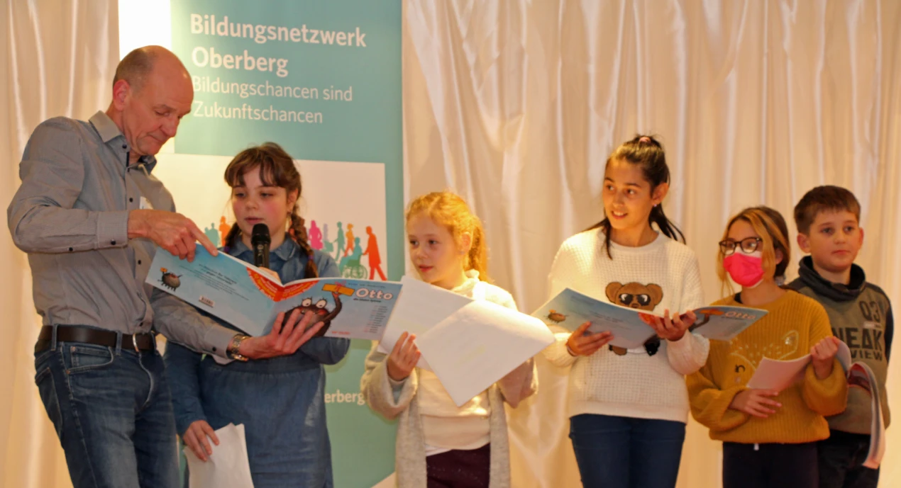 Norbert Fischer, pädagogischer Mitarbeiter des Bildungsbüro Oberberg unterstützte die mehrsprachig Vortragenden auf der Bühne (v.l.): Viktoria (deutsch), Melanie (russisch), Nadira (kroatisch), Anisa (englisch) und Dennis (ukrainisch) lasen in ihrer jeweiligen Muttersprache vor. (Foto: OBK) 