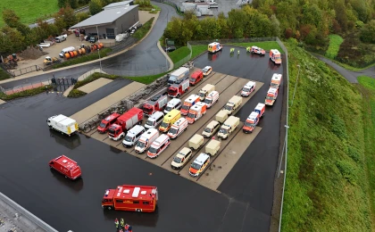 Drohnenaufnahme eines Parkplatzes mit einer Vielzahl von Einsatzfahrzeugen, darunter Rettungswagen, Feuerwehrfahrzeuge und andere Einsatzfahrzeuge. Im Hintergrund ein Gebäude, Grünflächen und Straßen.