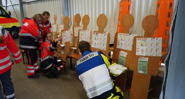 Personen in Rettungskleidung arbeiten mit hölzernen Figuren, die mit Dokumenten und Diagrammen versehen sind, in einem Innenraum mit medizinischen Geräten.