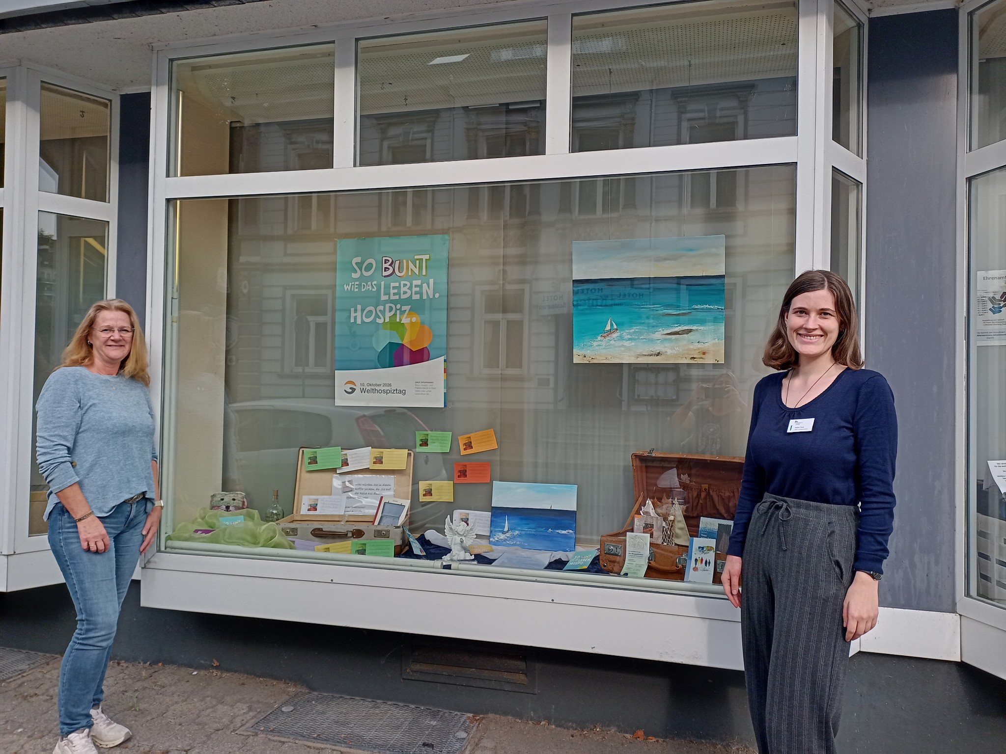 Zwei Frauen stehen vor einem Schaufenster mit einer Ausstellung. Im Fenster sind Poster, Bilder mit Meer-Motiven und verschiedene Gegenstände zu sehen.