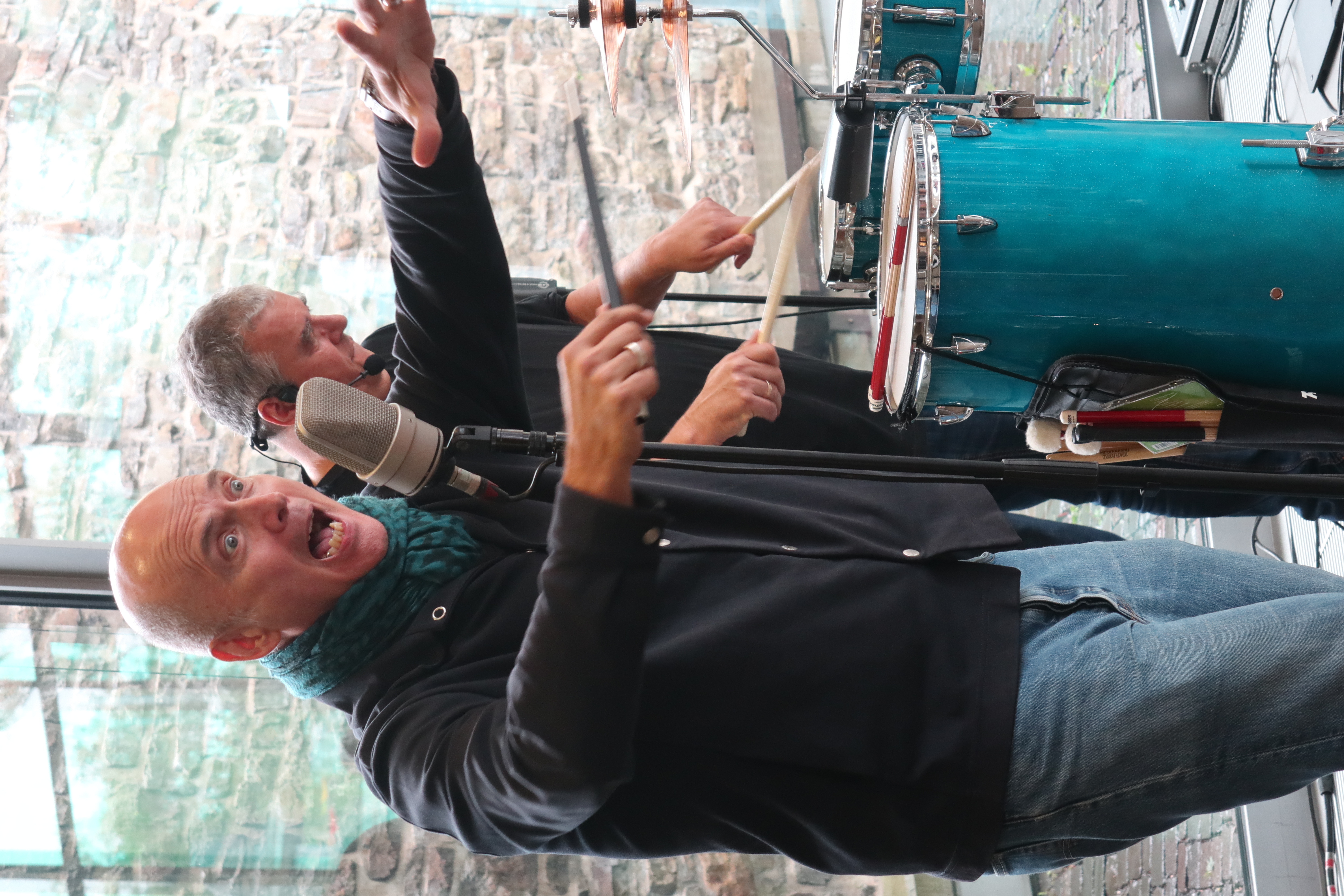 Ein Mann mit Glatze und Schal singt energisch in ein Mikrofon, während ein anderer Mann im Hintergrund Trommeln spielt. Im Hintergrund ist eine Steinwand und ein Fenster zu sehen.