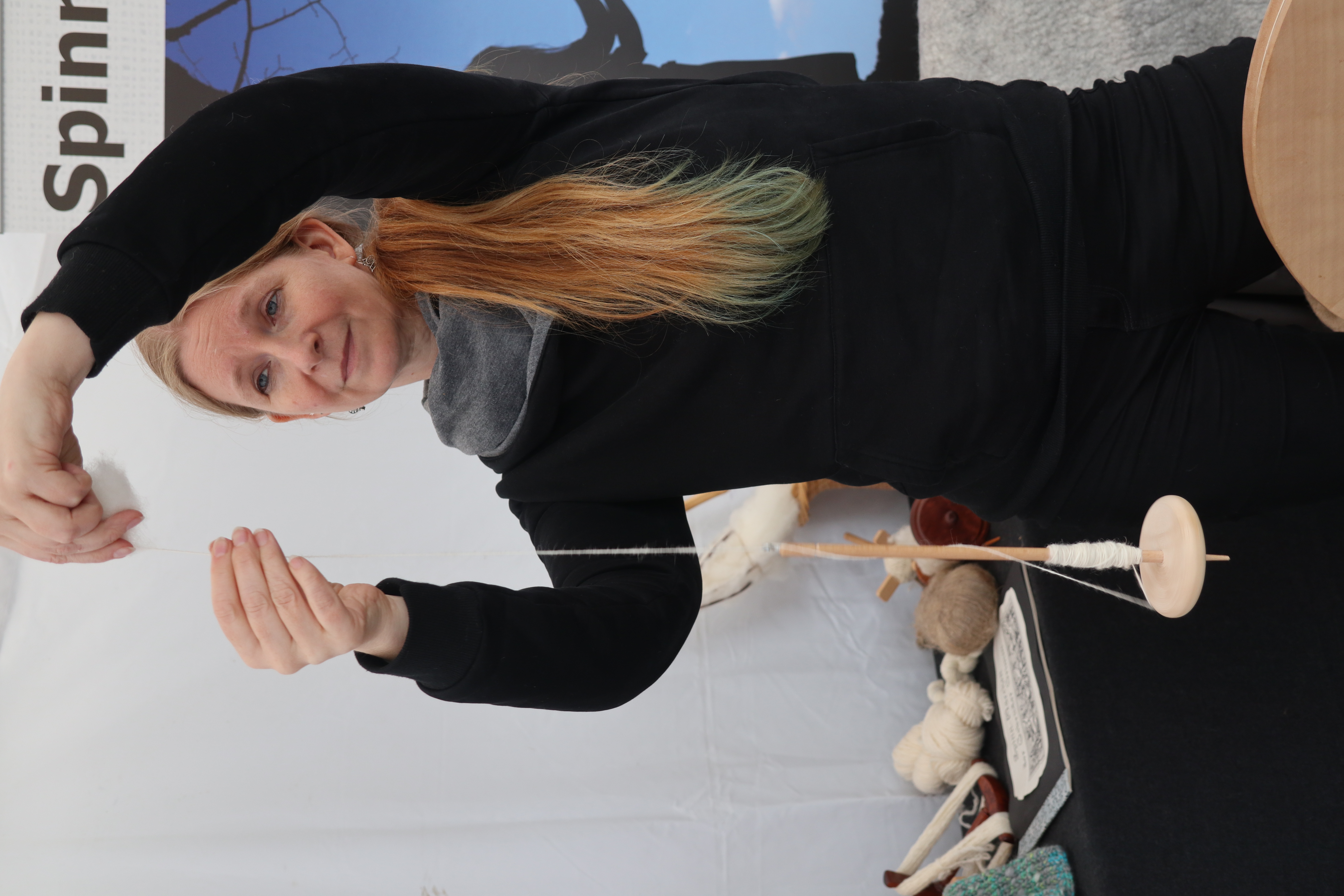 Eine Frau mit langen rötlichen Haaren hält in ihren Händen eine Handspindel und spinnt Wolle. Im Hintergrund sind Wollprodukte und ein Stand zu sehen.
