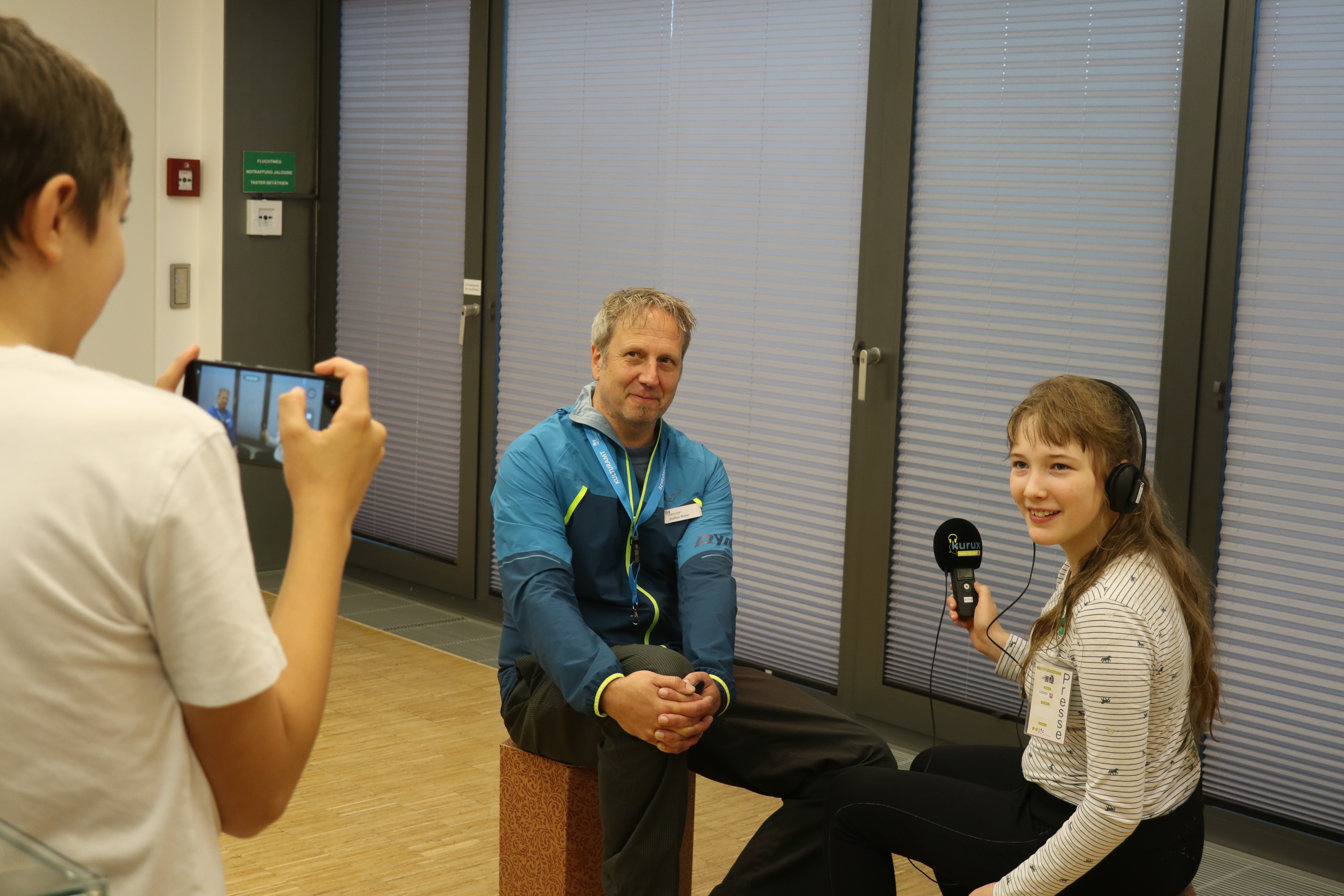 Eine junge Person mit Headset und Mikrofon interviewt einen Mann in blauer Jacke, während eine dritte Person das Geschehen mit einem Smartphone fotografiert. Im Hintergrund sind Fenster mit Jalousien zu sehen.