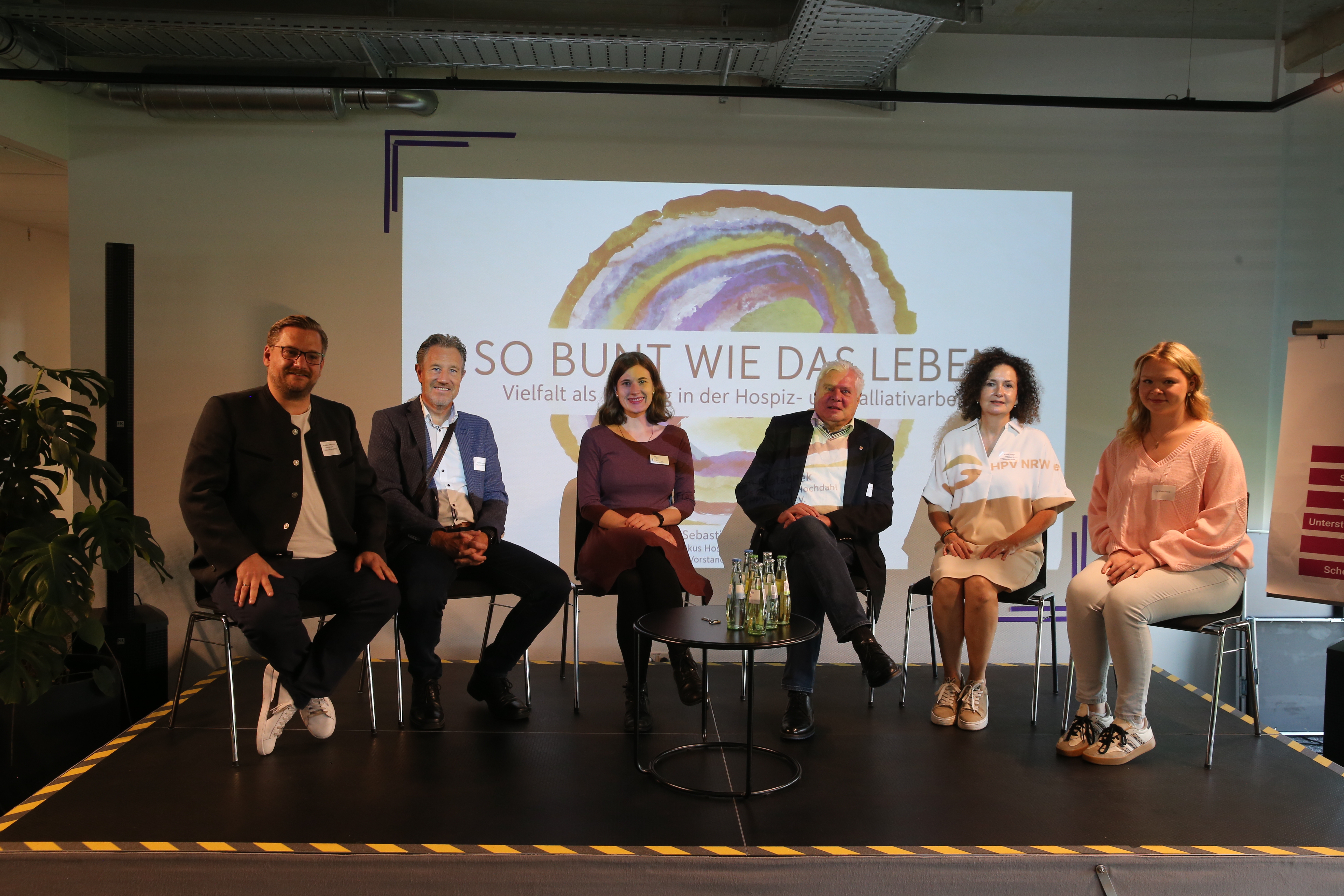 Sechs Personen sitzen nebeneinander auf einer Bühne vor einer Leinwand mit dem Titel 'So bunt wie das Leben: Vielfalt als Chance in der Hospiz- und Palliativarbeit'. Vor ihnen steht ein kleiner Tisch mit Wasserflaschen.