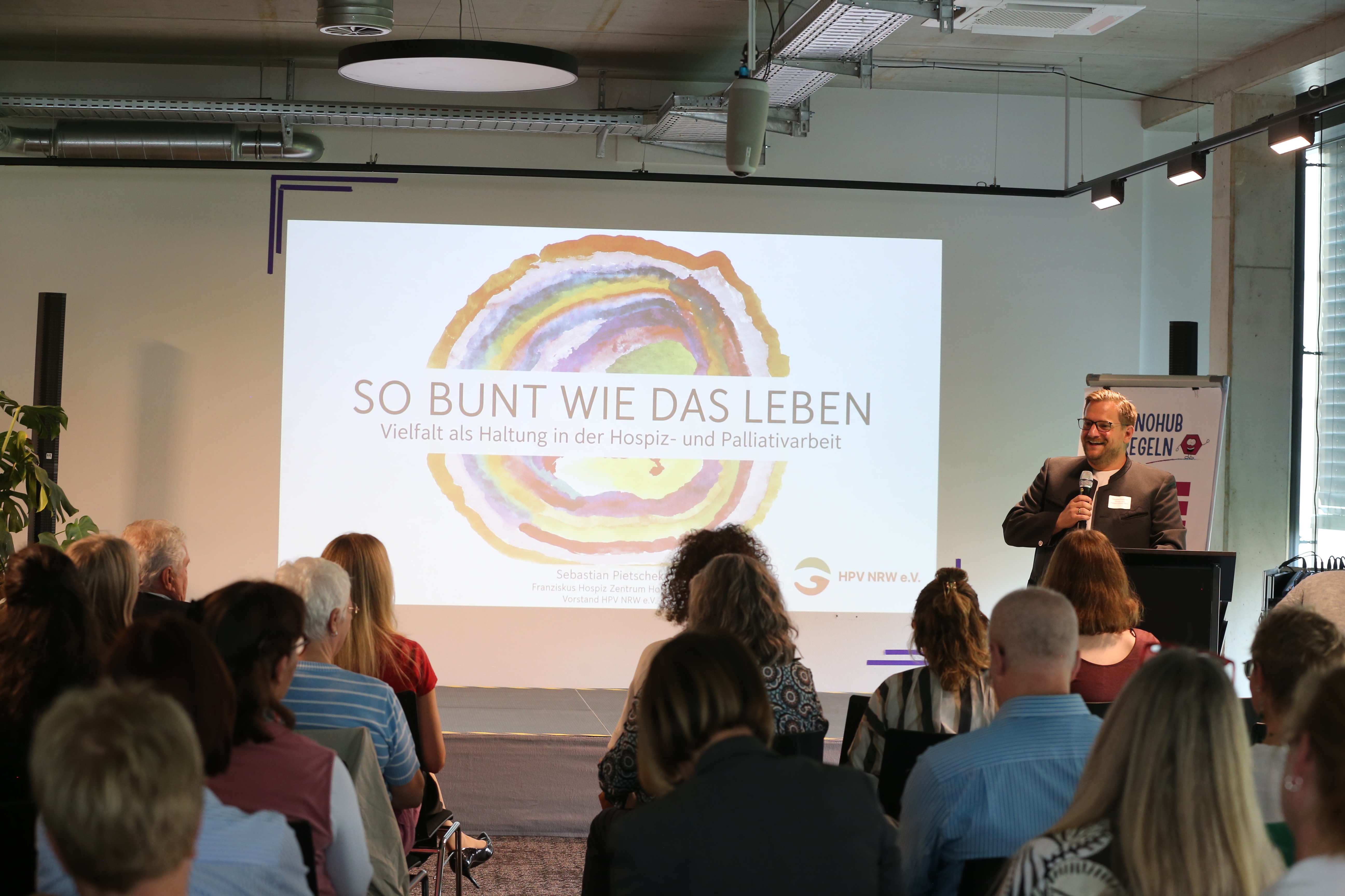Ein Mann hält eine Präsentation vor Publikum in einem Konferenzraum. Auf der Leinwand steht 'So bunt wie das Leben - Vielfalt als Haltung in der Hospiz- und Palliativarbeit'.