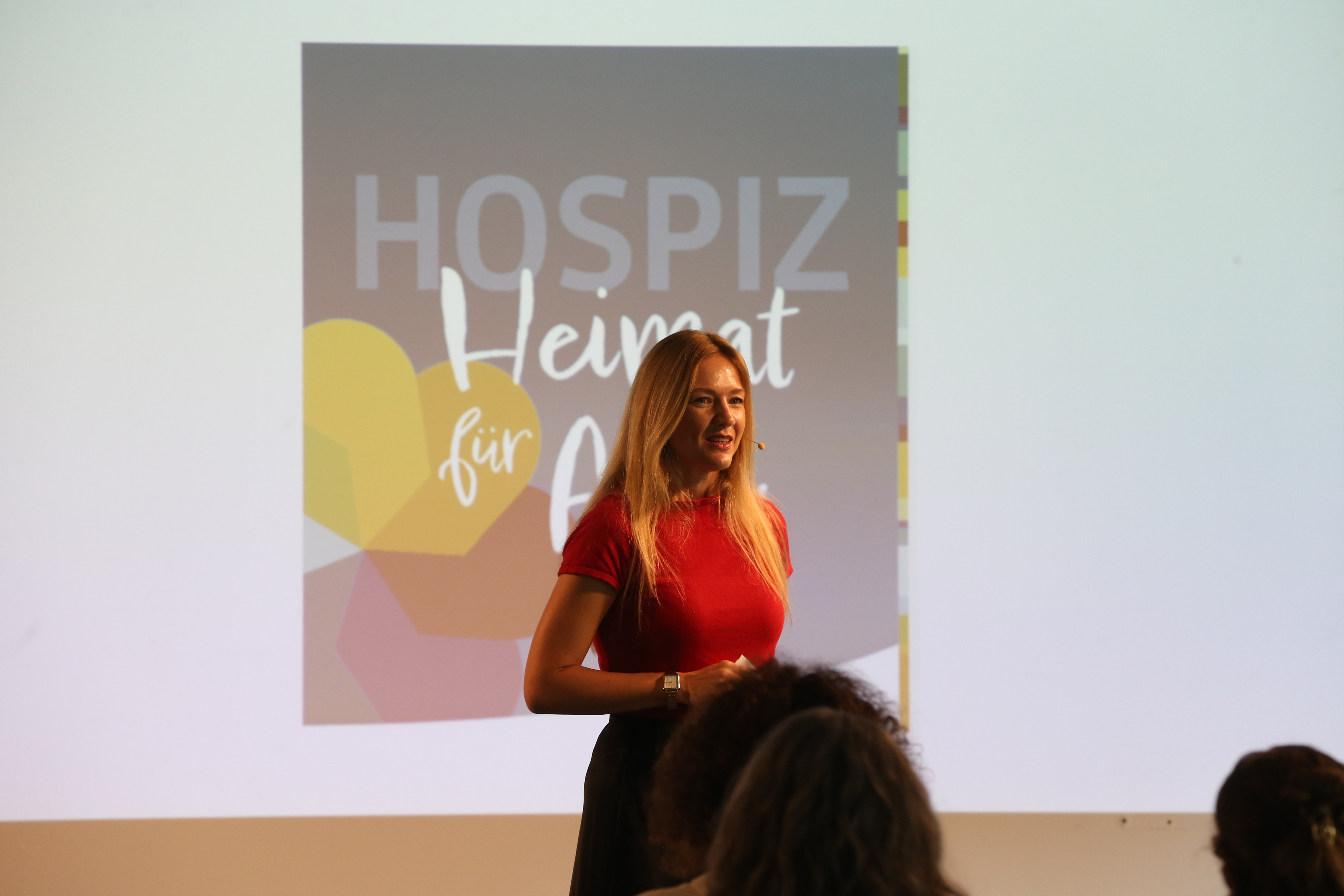 Eine Frau mit langem blondem Haar und rotem Oberteil spricht vor einem Publikum. Im Hintergrund ist ein Plakat mit der Aufschrift 'Hospiz Heimat für Alle' und Herzgrafik sichtbar.