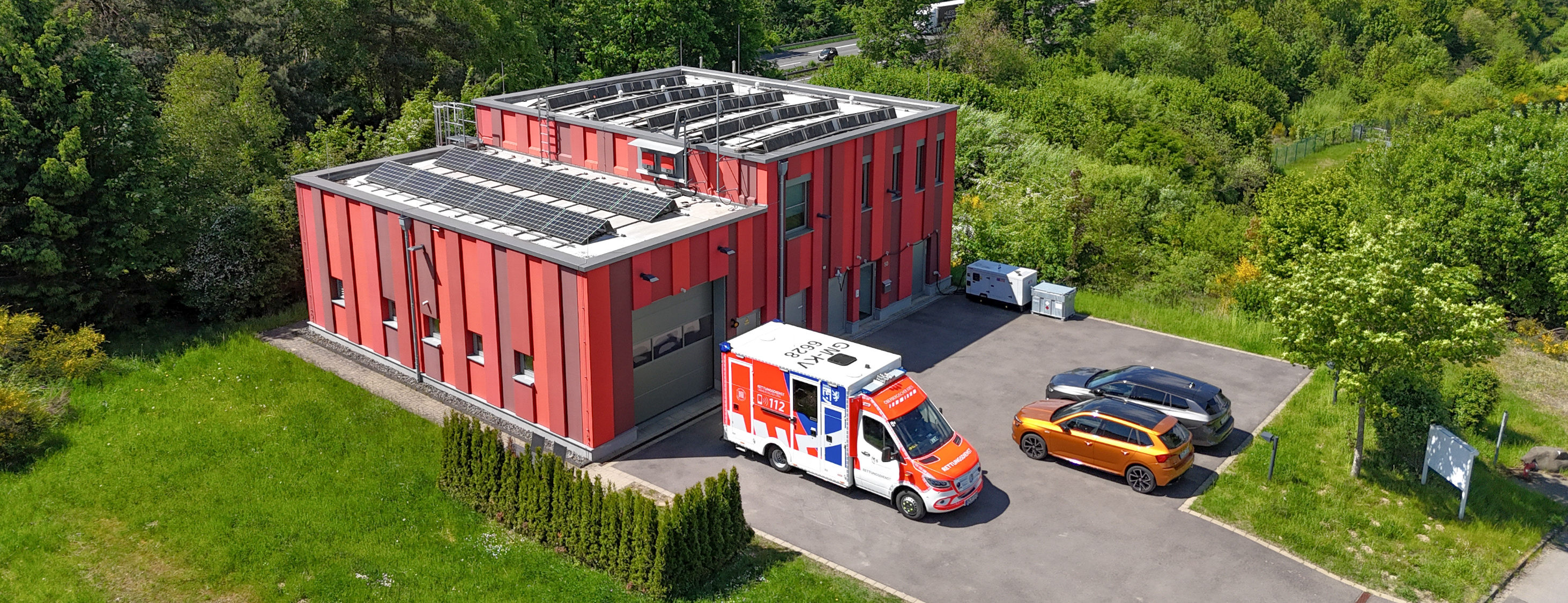 Ein modernes, rotes Gebäude mit Solarpaneelen auf dem Dach, umgeben von Bäumen. Auf dem Parkplatz stehen ein Rettungswagen und zwei Autos.